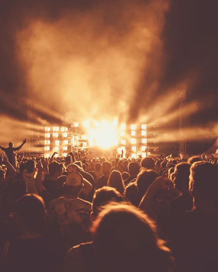 Festival audience facing a stage of bright tungsten beams under a haze-lit sky.