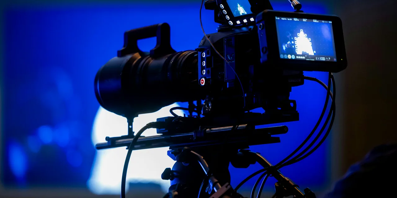 Cinema broadcast camera with field monitor and lens on a tripod under blue rim light.