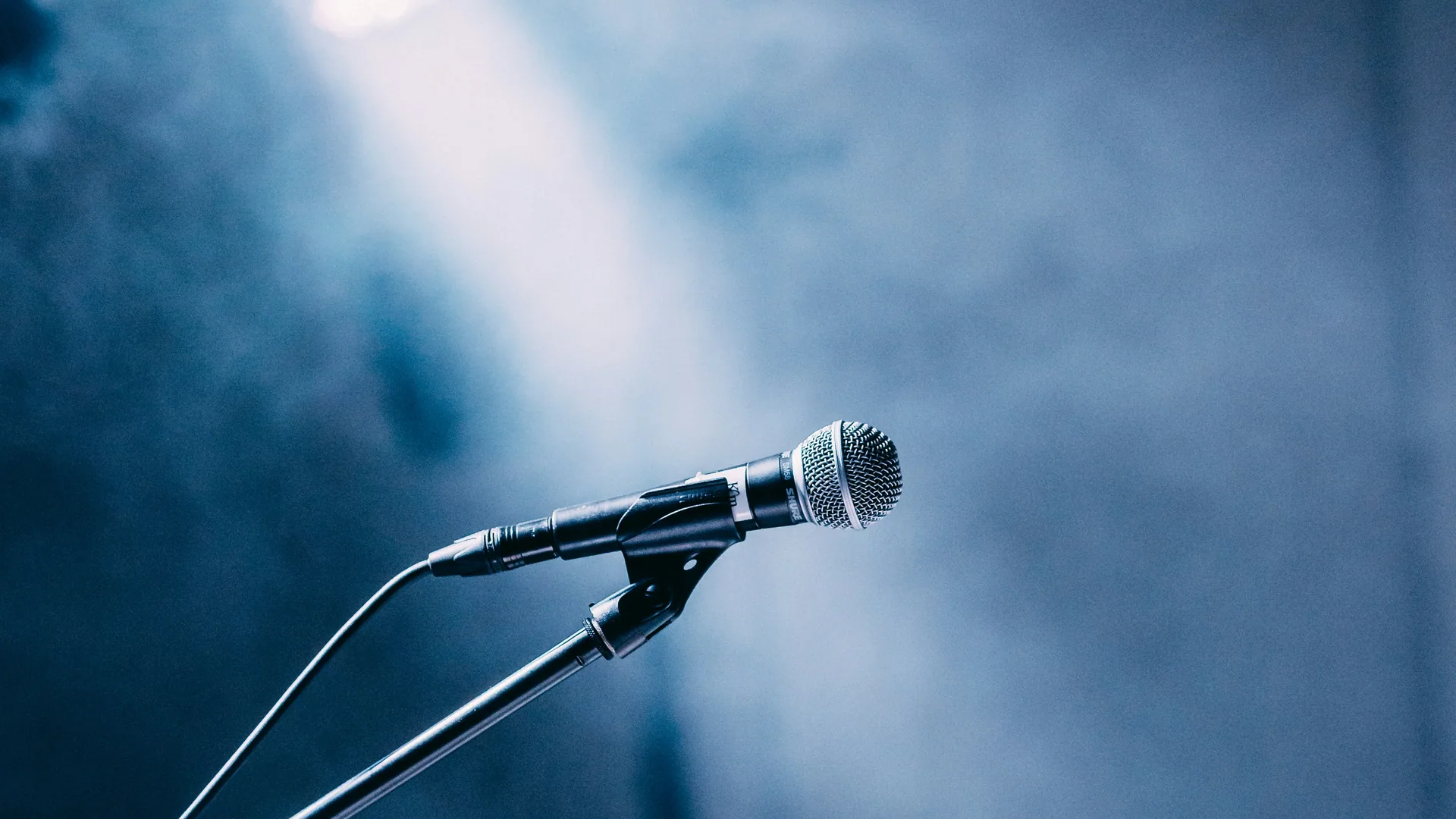 Vocal microphone on a stand spotlit by a single beam against a haze-filled stage.