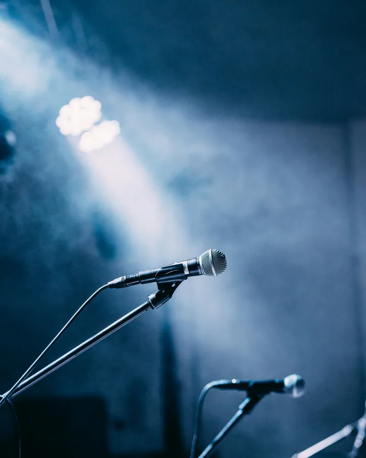 Vocal microphone on a stand spotlit by a single beam against a haze-filled stage.