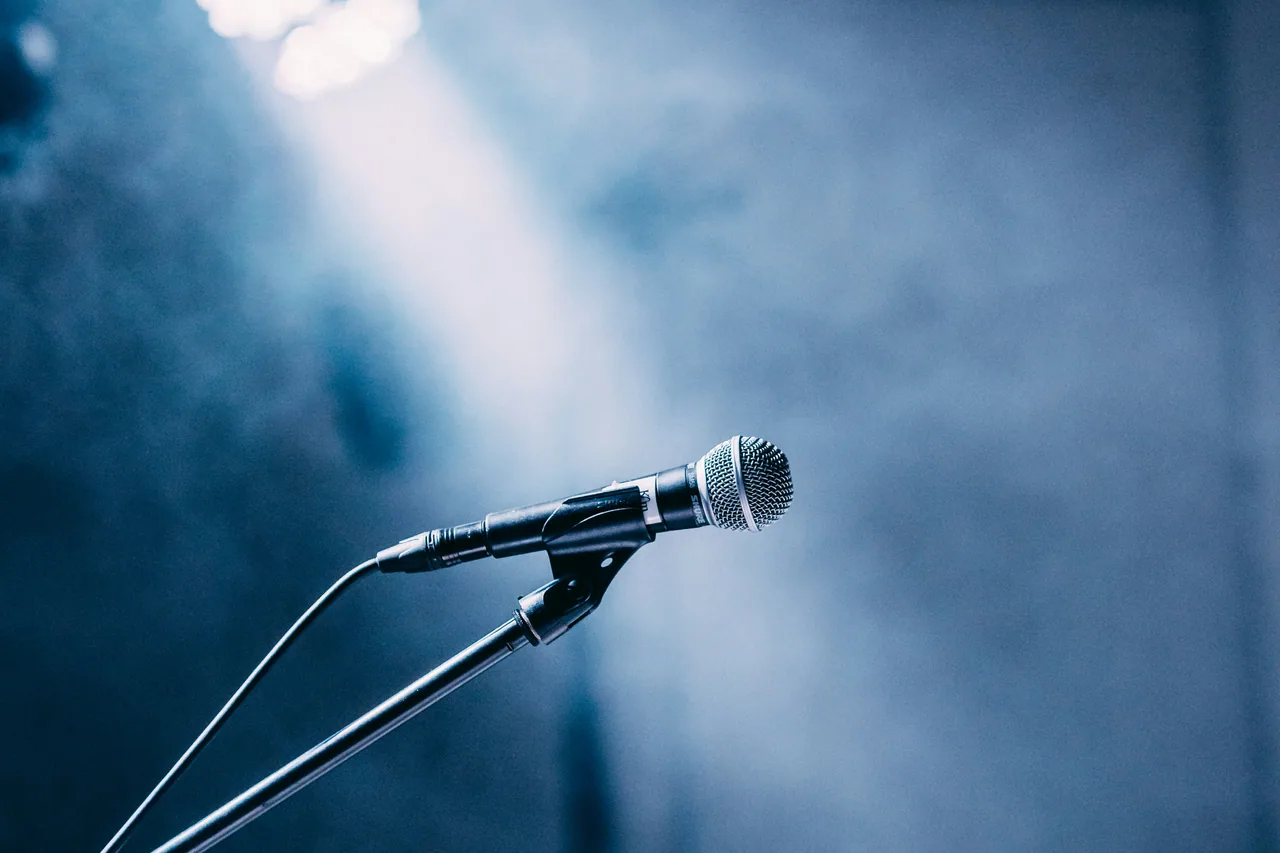 Vocal microphone on a stand spotlit by a single beam against a haze-filled stage.