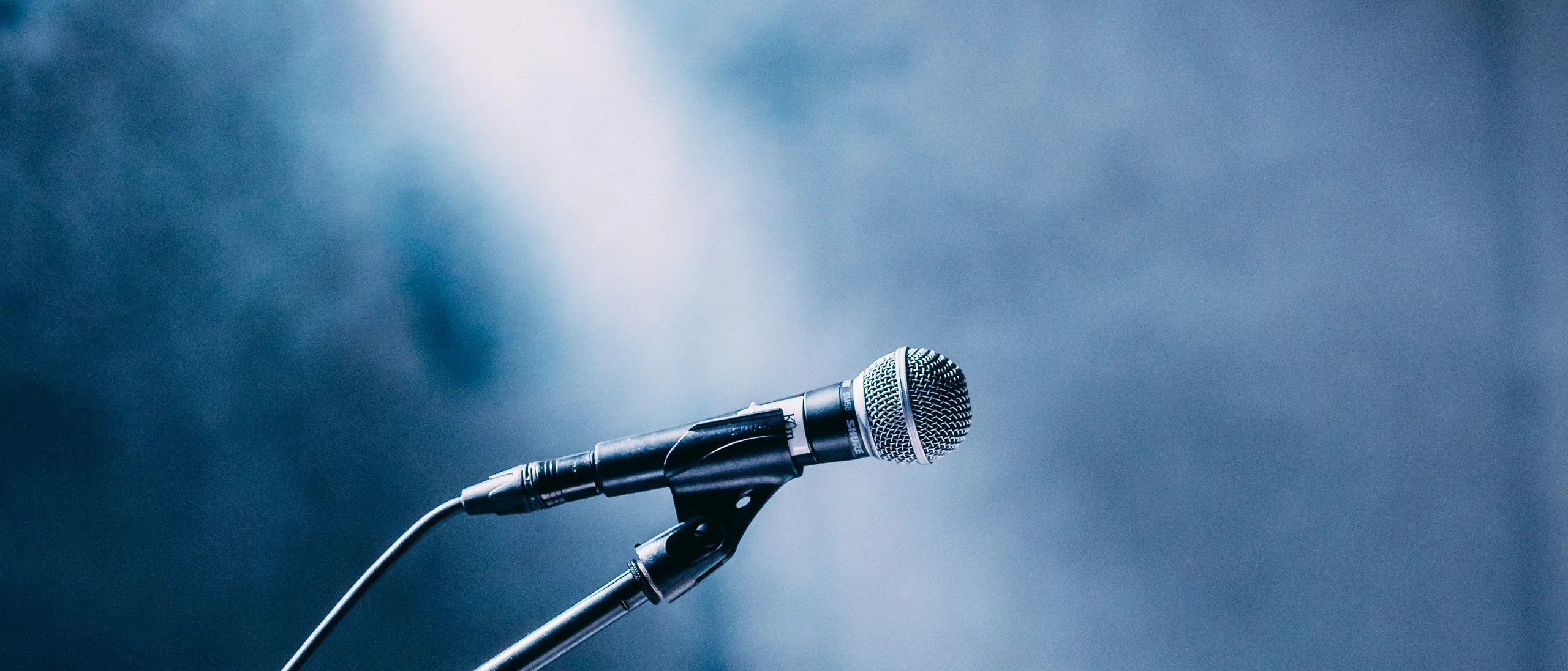 Vocal microphone on a stand spotlit by a single beam against a haze-filled stage.