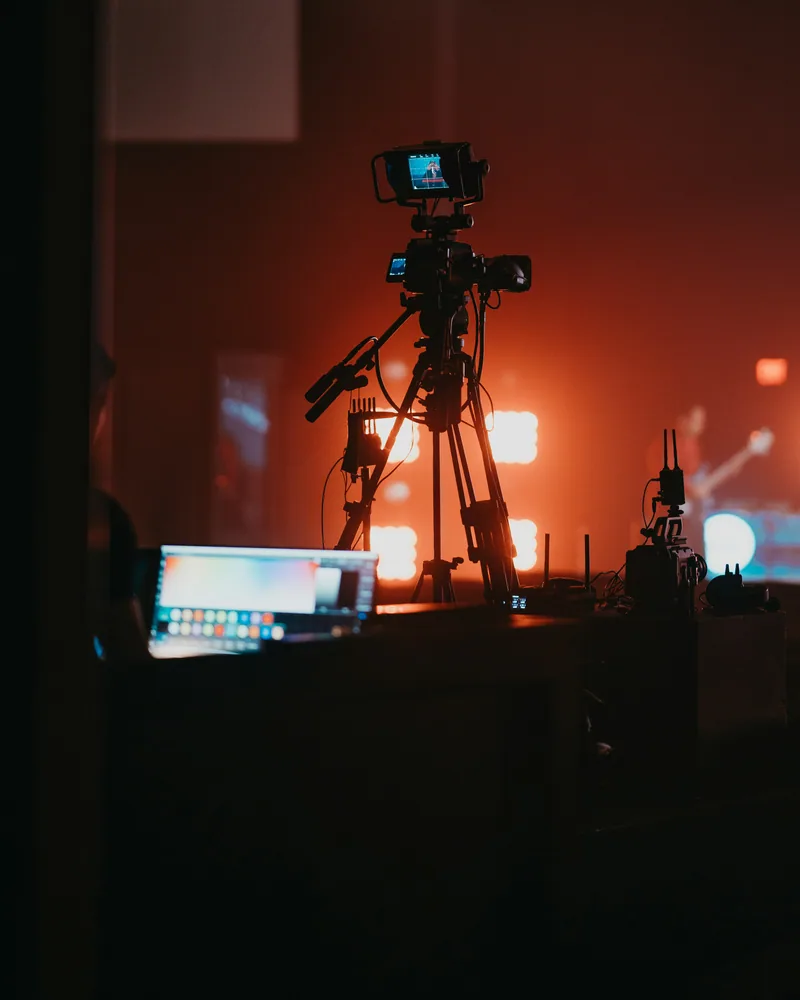 Broadcast camera on a tripod with viewfinder backlit by stage lighting.