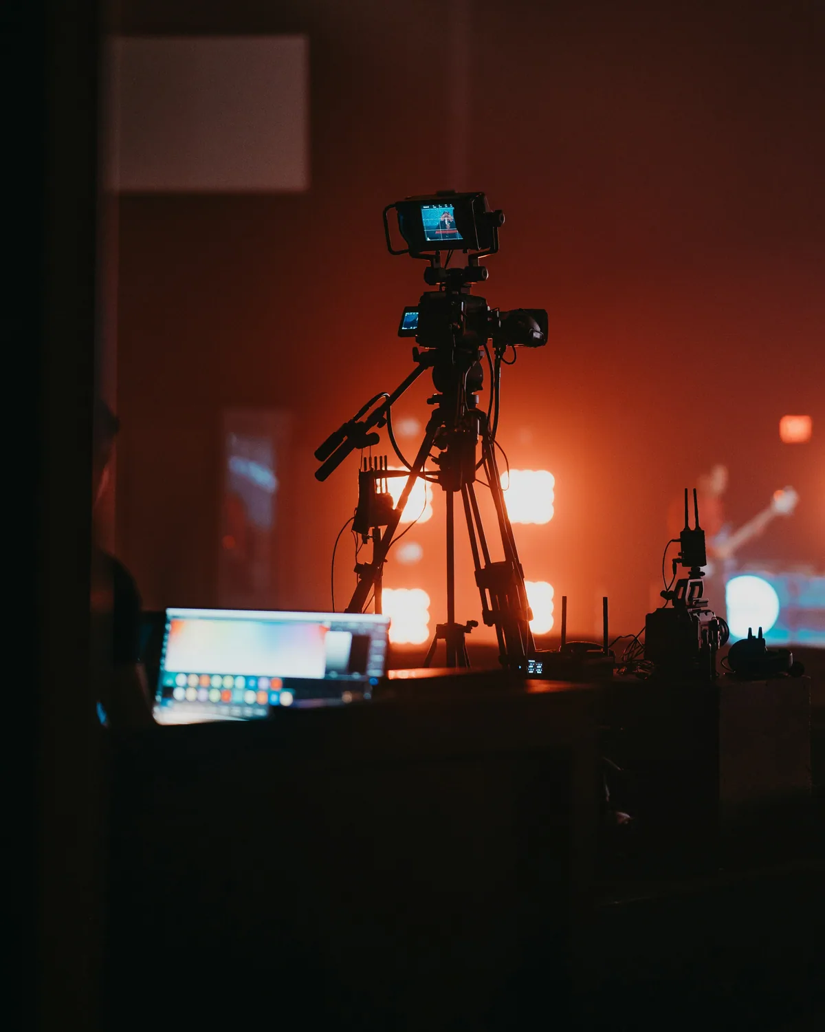 Broadcast camera on a tripod with viewfinder backlit by stage lighting.